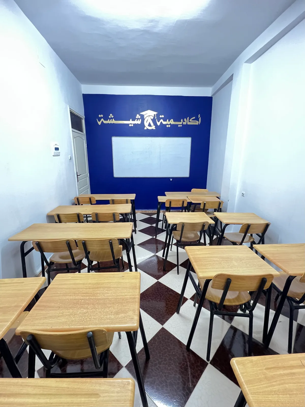 قاعة دراسة بجدار أزرق يحمل اسم أكاديمية شيشة بالذهبي فوق سبورة بيضاء، وطاولات خشبية بأرجل سوداء